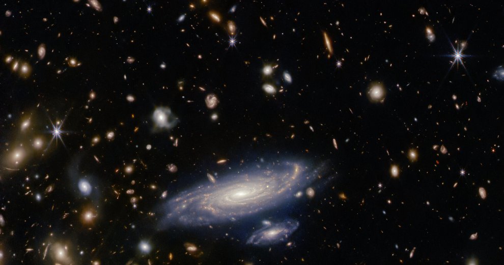 A detailed spiral galaxy against a crowded field of galaxies interspersed with bright 8-pointed stars on a dark background.