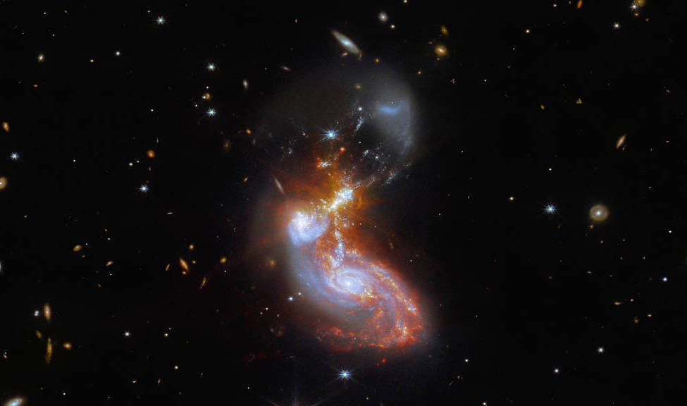 Two merging galaxies form a white and red bean-like shape against a black sky. The cores of the galaxies are colored blue, and one is above and to the left of the other.