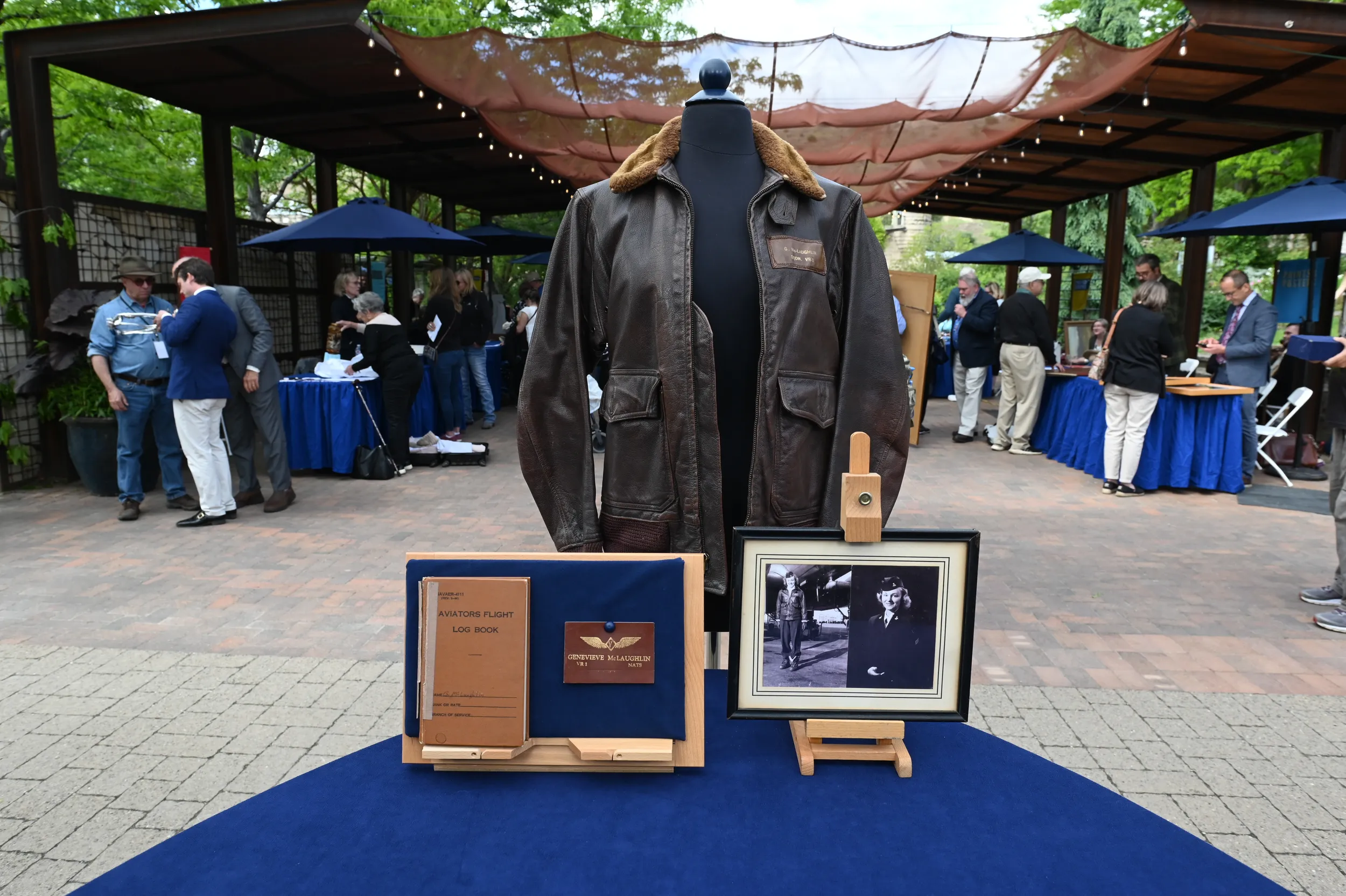 A brown leather woman's jacket sits on an appraisal table with a framed photograph of a blonde woman wearing the jacket. Next to the picture is an aviation log book