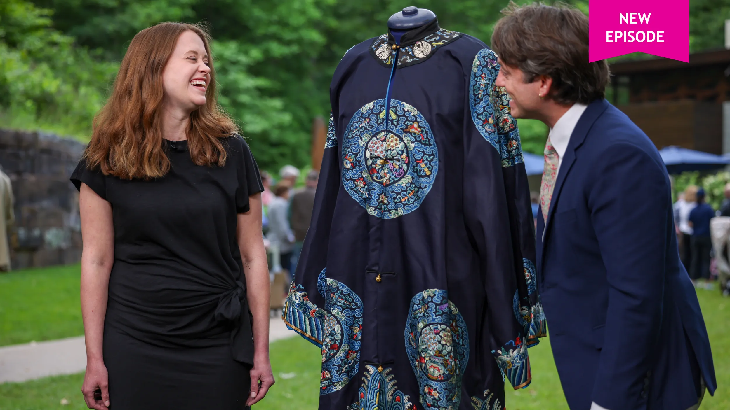 A young female guest with red hair laughs at something an appraiser says. Between them is a navy blue silk Chinese robe with red and light blue stripes on the bottom and sleeves.