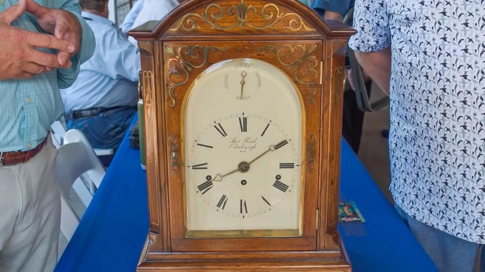Close up of a wooden clock with a cream clock face placed on a blue table with two people standing on either side.