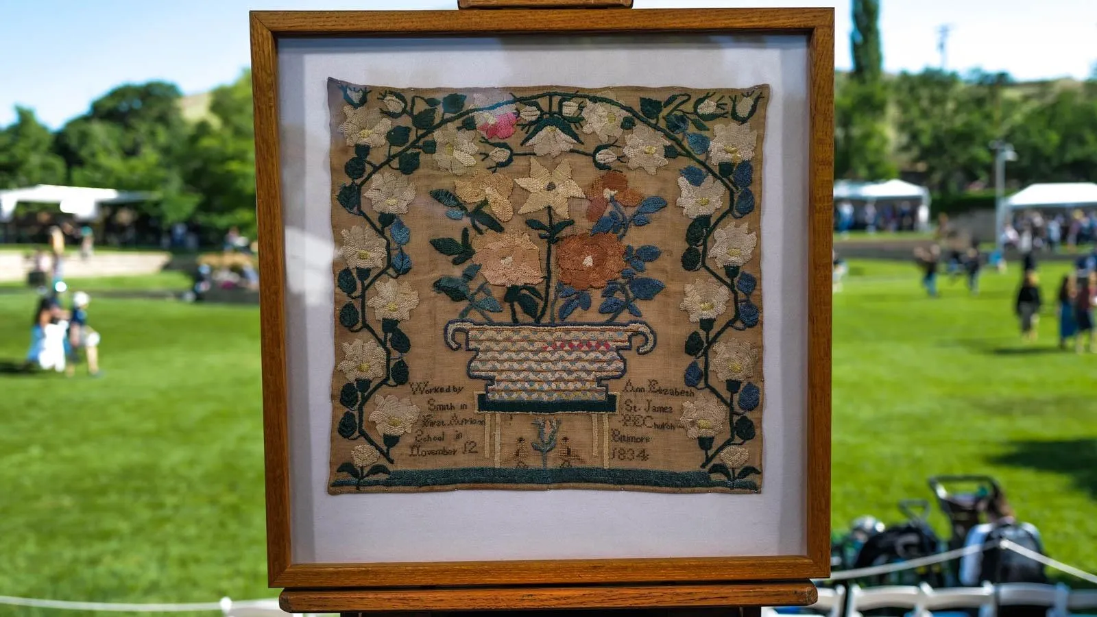 A framed needle-work sampler with a vine of white and blue flowers cascading up the left of the piece, across, and down the right. In the center are orange, yellow and blue flowers.