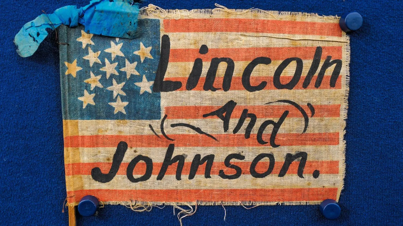 A close up on a small american flag with the words "Lincoln and Johnson" written in a black font. Above the stars on the flag on the left is a blue ribbon bow.