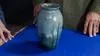 Blue Vase with forest like details against a blue background with three hands positioned on each side.