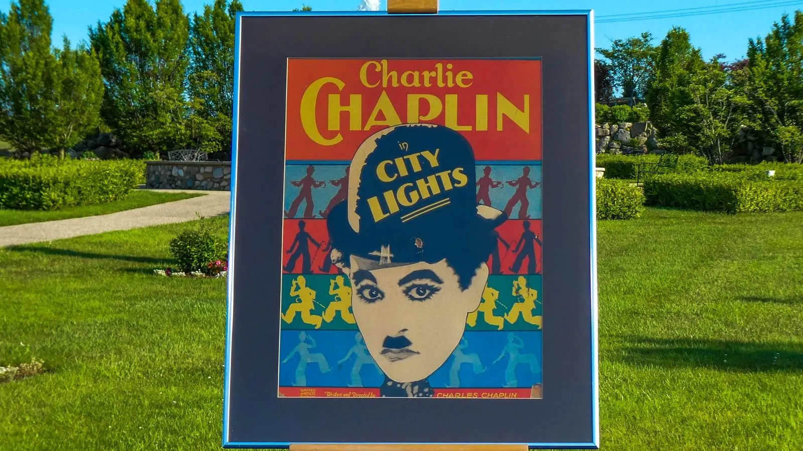 A close up poster for Charlie Chaplin's City Lights featuring his face in the foreground as black and white with a sillouette background in primary colors