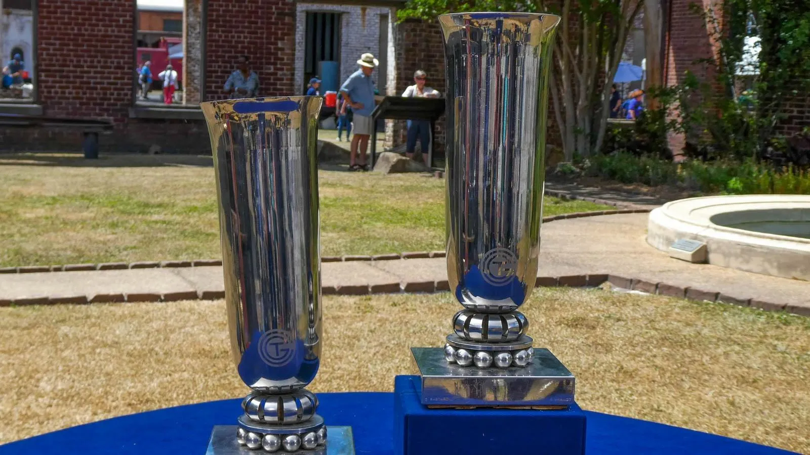 Two silver chrome cylindrical vases with square bases are placed on top of a blue table outside of a brick building.