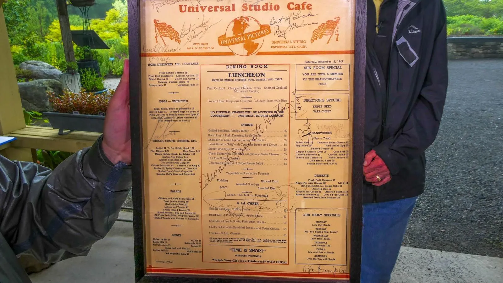 Two people hold up a larged framed menu labelled "Universal Studio Cafe" signed with many signatures.