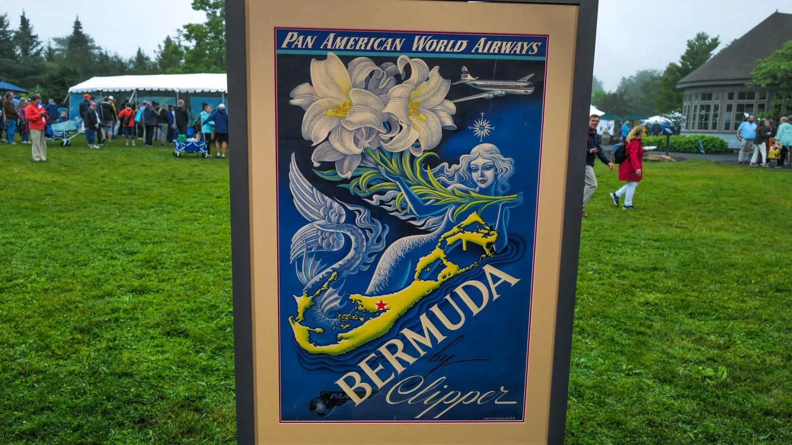 A poster is in a dark grey frame. The poster says bermuda clipper in white letters with a green outline of the country above it. On top of Bermuda is a mermaid with white flowers.