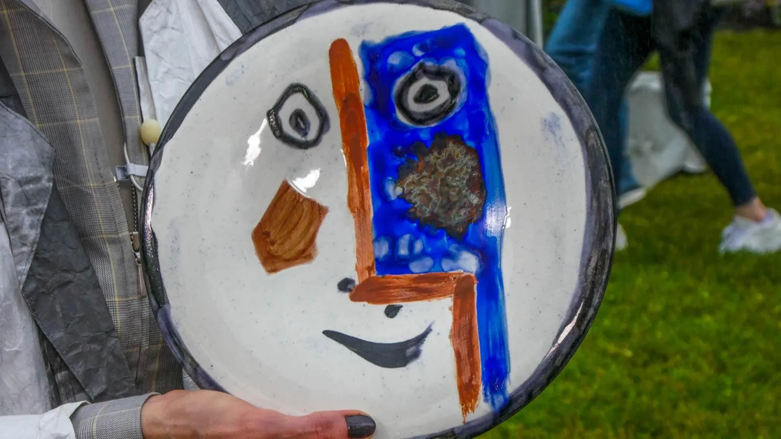 A hand holding a plate with a cubist face on it in colors of brown and blue against a white background.