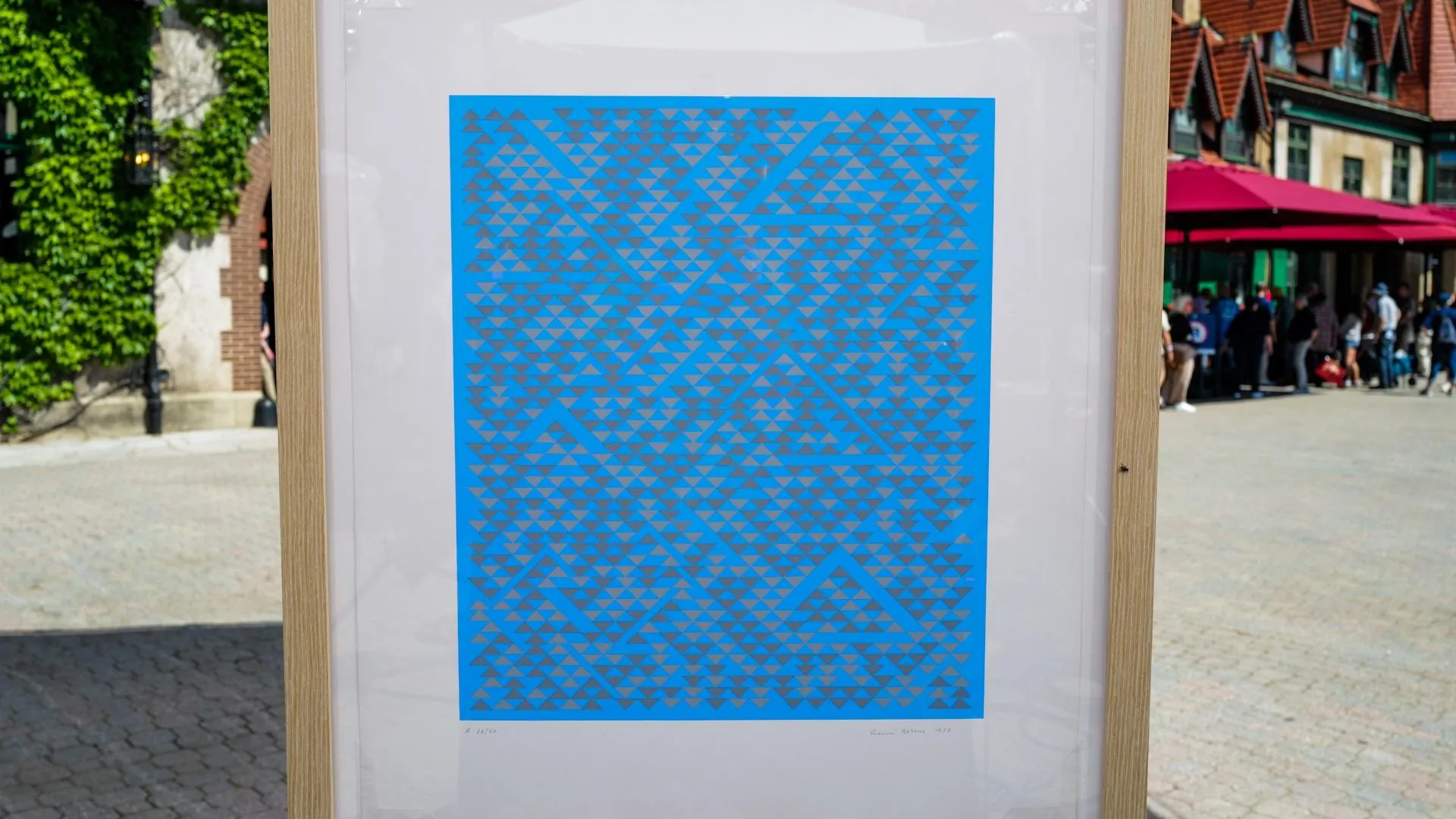 A blue screenprint with a small trianular pattern is centered in a wooden frame placed on an easel.