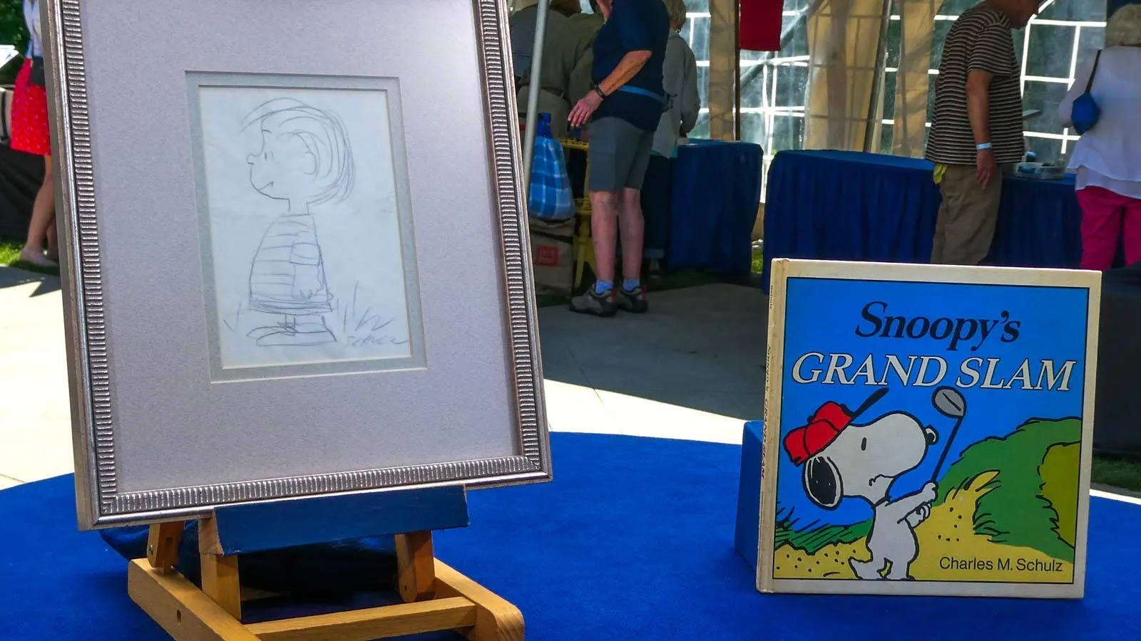 A framed pencil sketch of Linus from the peanuts comic series next to a book called "snoopy's grand slam" featuring snoopy hitting a golf ball.