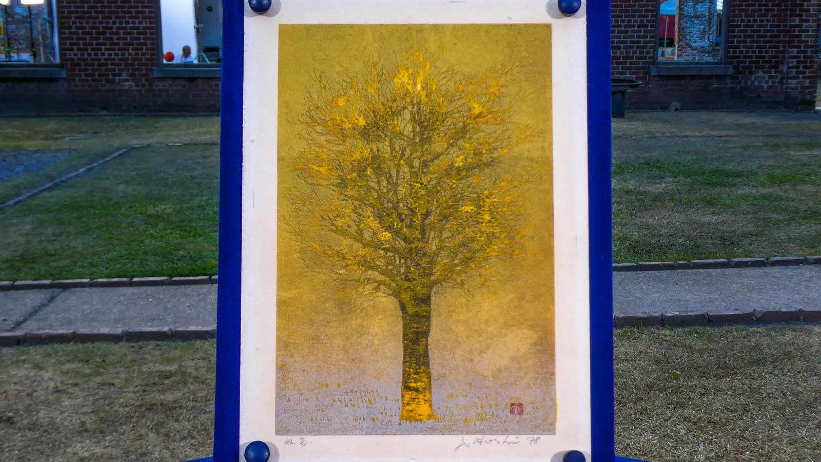 A woodblock print of a tree with a yellow tint mounted on a blue  poster board