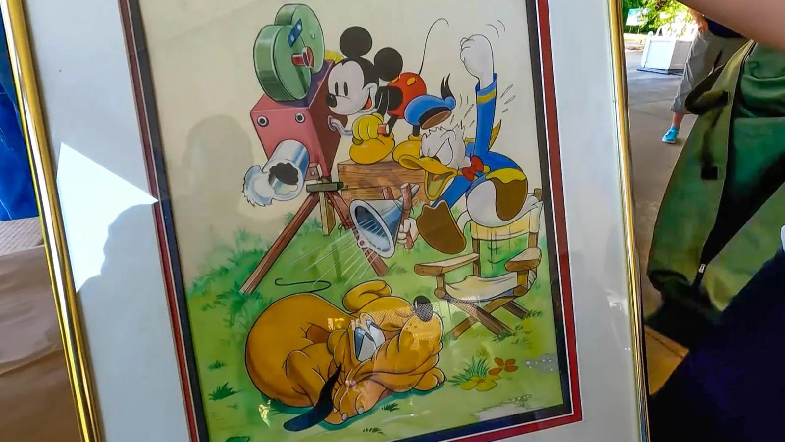 A framed image of Mickey Mouse, Donald Duck, and Pluto with an old movie camera being held up by a man in blue.