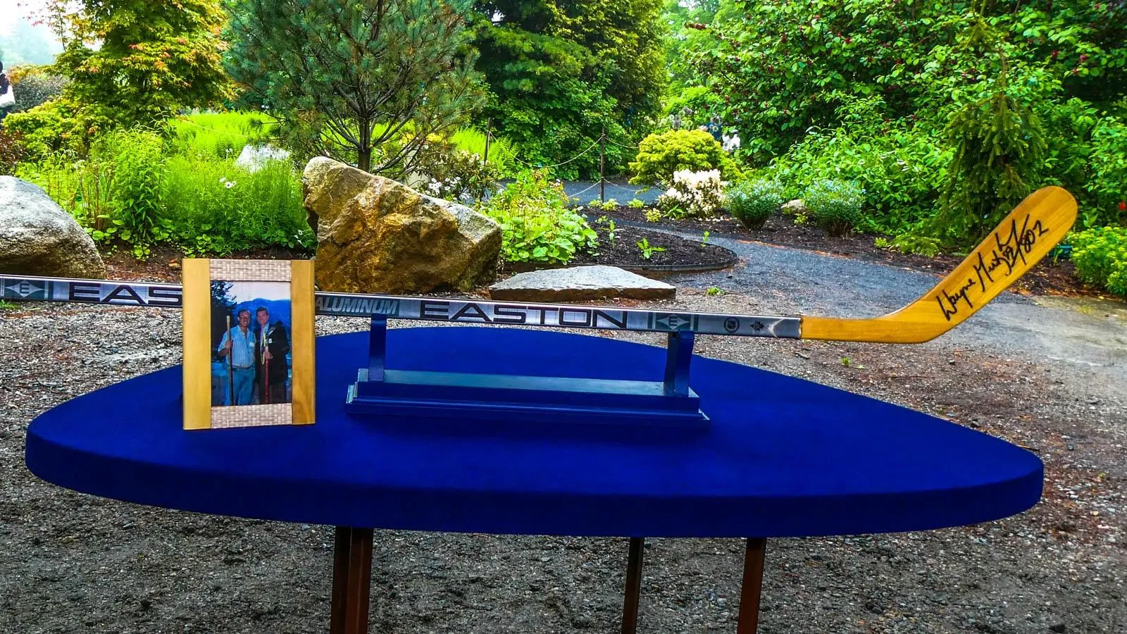 A signed Easton hockey stick with a wooden blade sites on a circular blue table next to a photo of two men standing side by side.