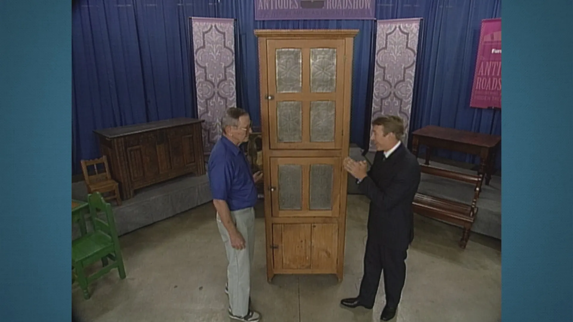 Leigh Keno (right) and a male guest, both standing, discuss an antique pie safe.
