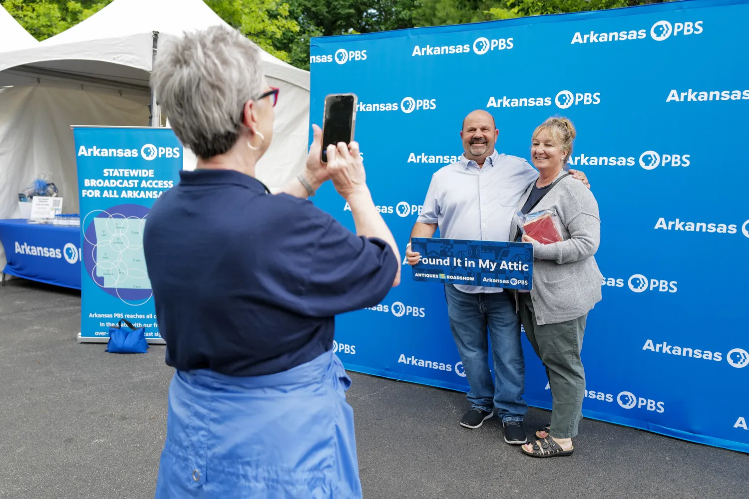 Two guests smile in front of an Arkansas PBS banner, their photo being taken by a ROADSHOW event volunteer.