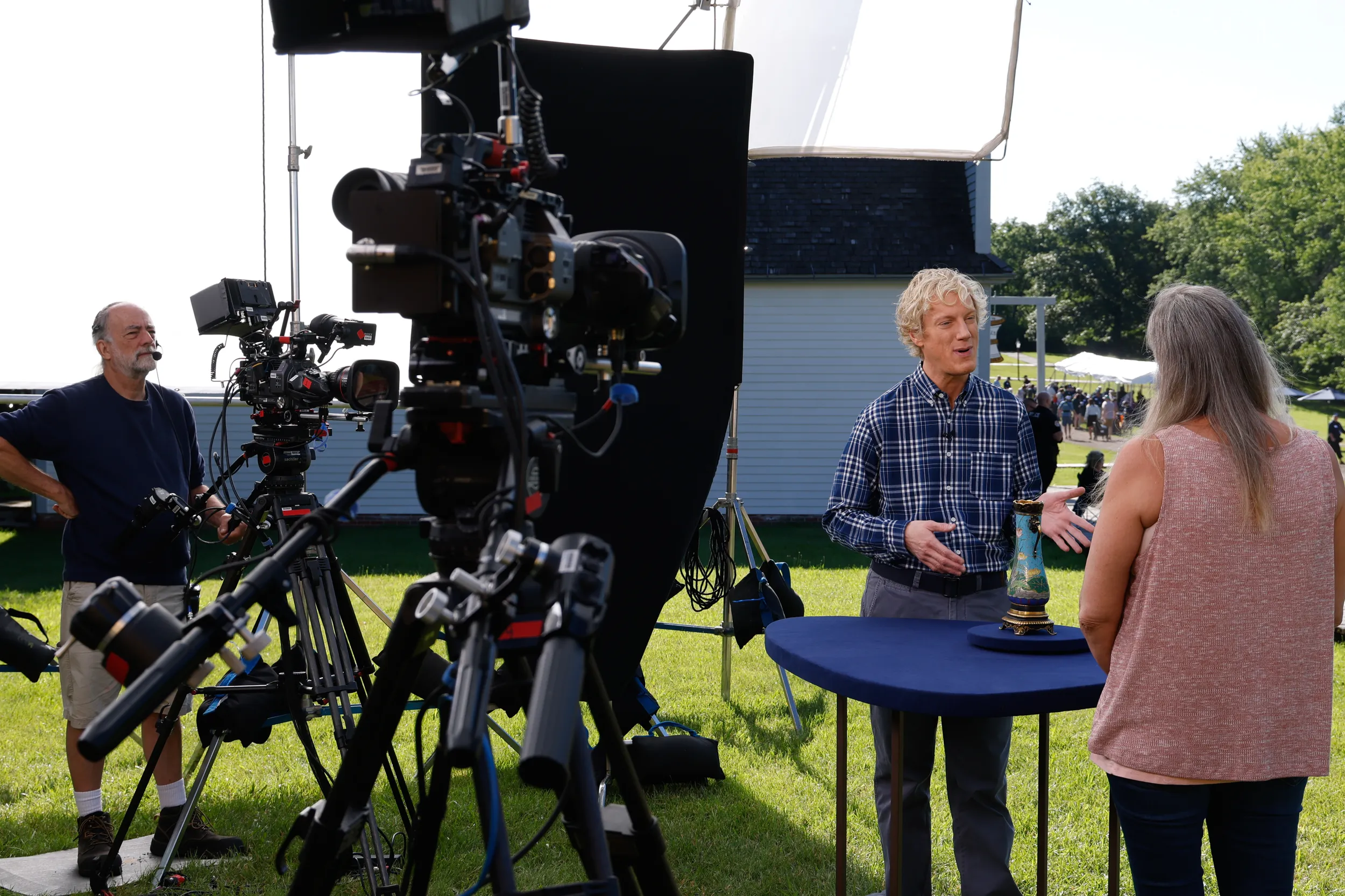 A young appriaser in a checkered blue shirt speaks to the female guest as he tells her about her blue vase. A camera records the appraisal as a camera operator watches from a monitor.