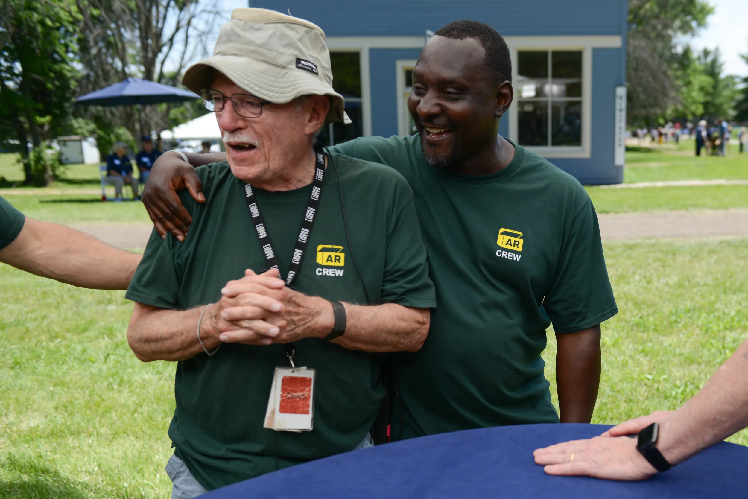 Two Roadshow crew members in green crew shirts clap each other on the back and laugh at something off screen as they stand next to an appraisal table.