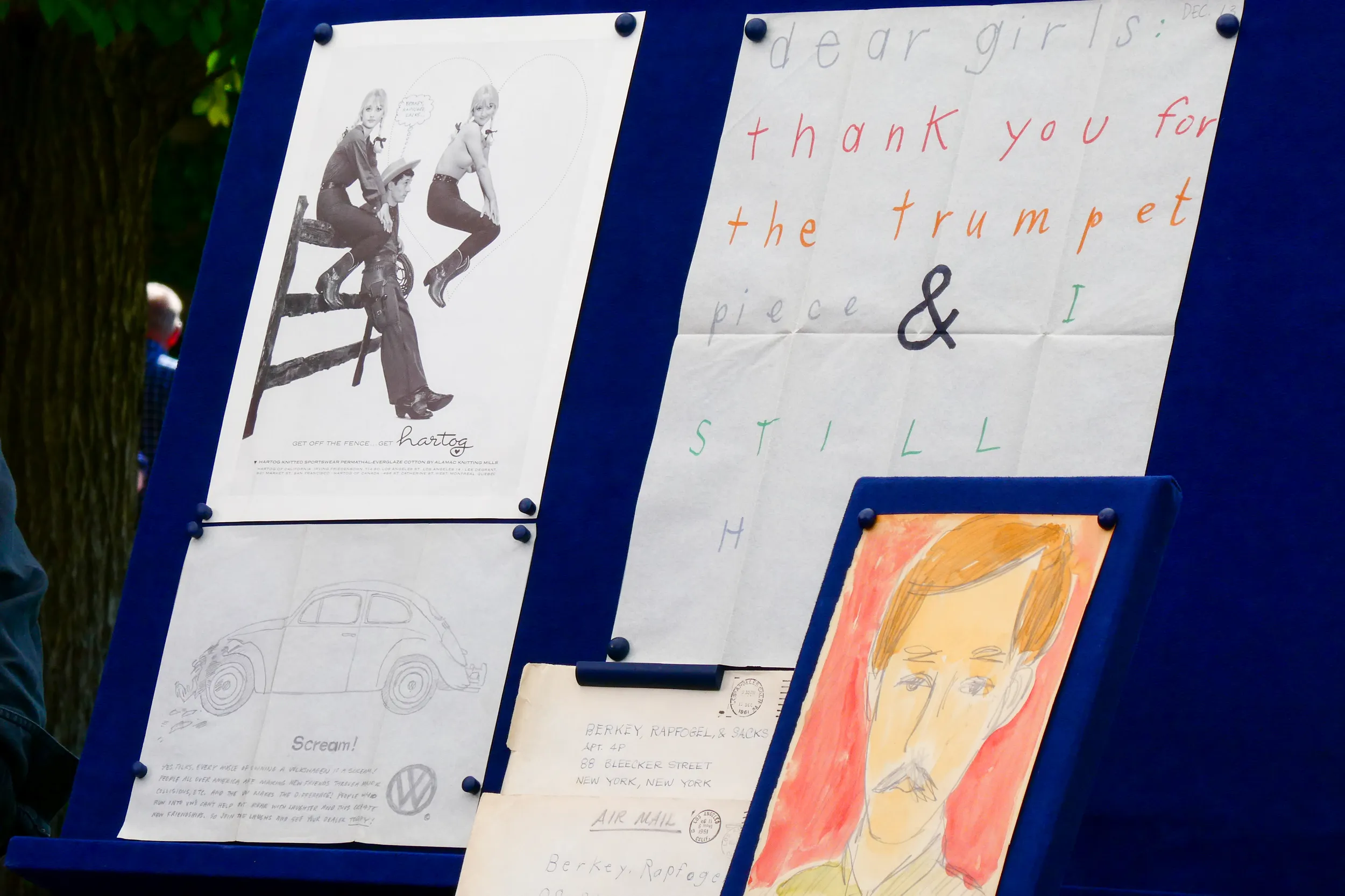Collection of drawings and sketches tacked to a blue board including a drawing of a volkswagen beetle car, a colored drawing of a man with a mustache and button down shirt, and a black-and-white ad