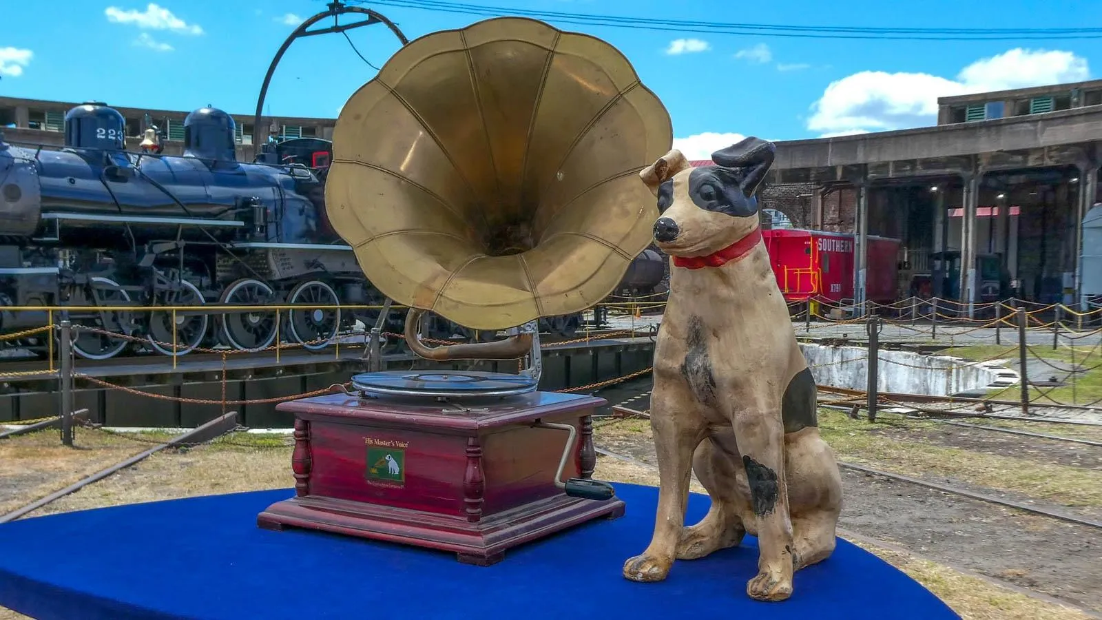 A gramophone record player with a maroon base sits on a table next to a dog statue against an outdoor background with a black train