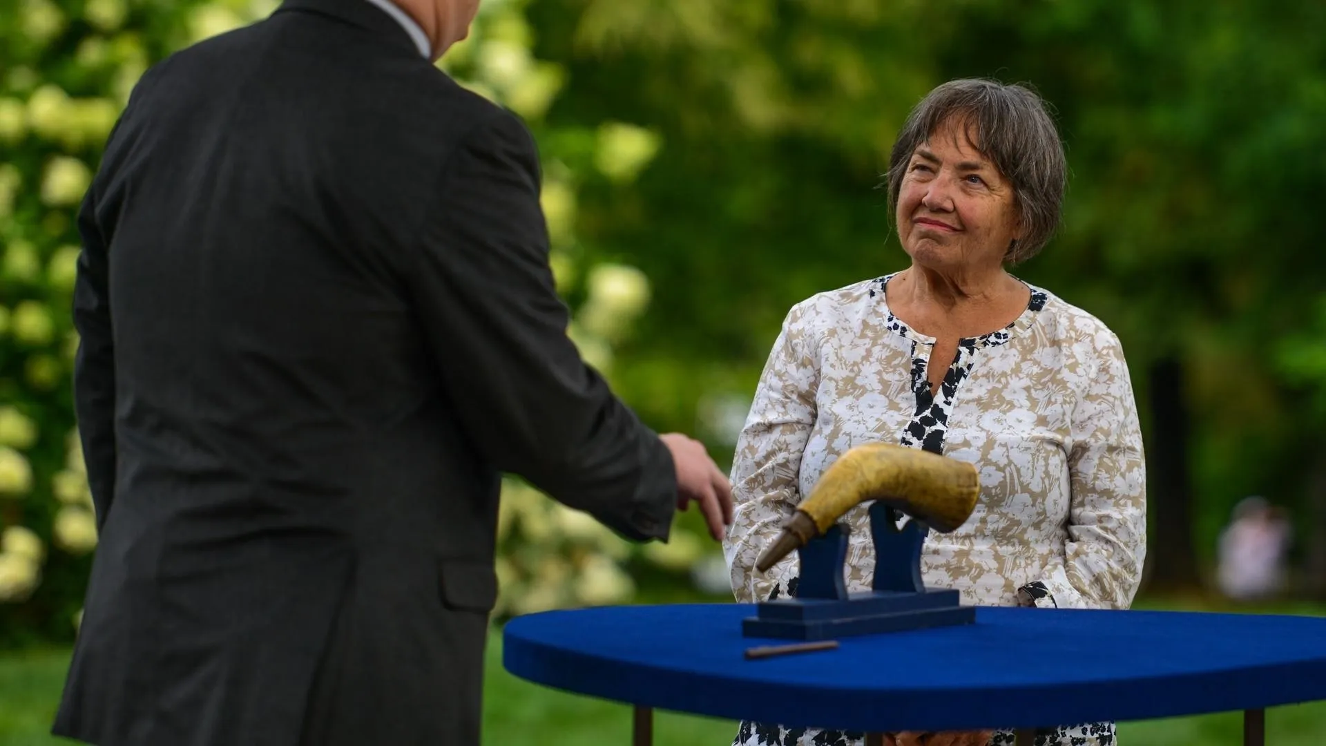 Women listening to folk art appraiser discussing a carved powder horn in front of shrubbery