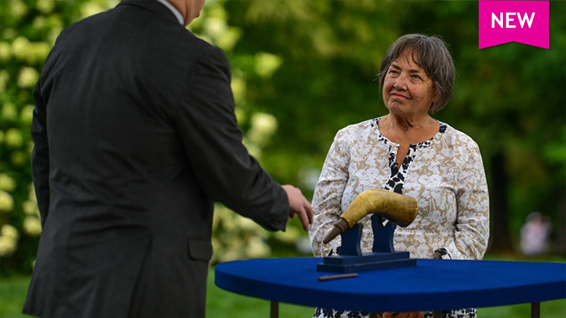 Women listening to folk art appraiser discussing a carved powder horn in front of shrubbery