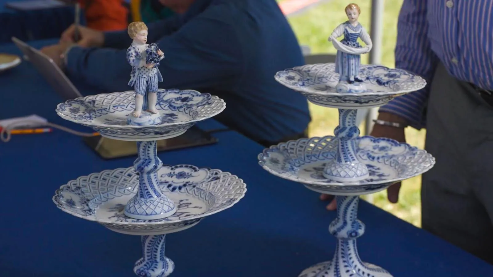 Two two-tiered dish stands in blue and white porcelain. On top of each tower is a statue of a child, one of a girl in a dress and towel and another of a boy holding grapes.