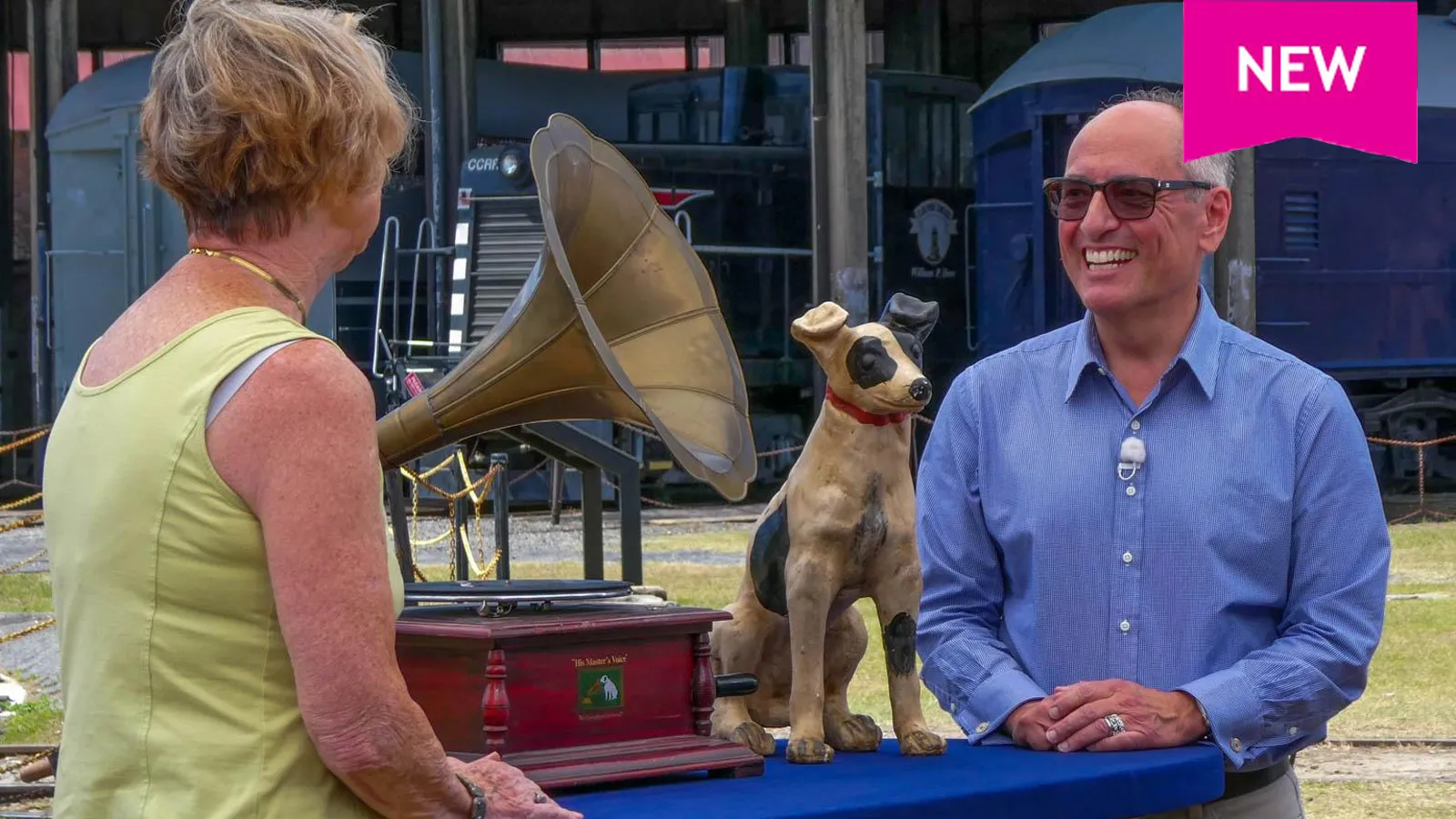 An appraiser in sunglasses faces the camera and smiles at a guest, whose bak is to us. There is a gramophone and a small dog statue on the appraisal table between them.