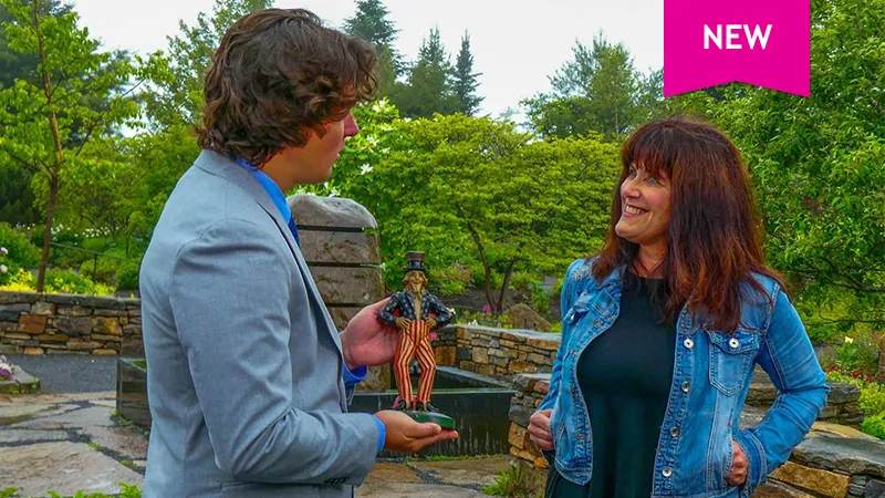 color photo of lady discussing an antique toy with appraiser Michael Bertoia on a patio with trees in the background