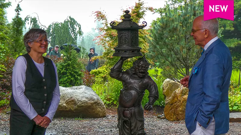 color photo of lady discussing an antique Chinese sculpture with appraiser Lark Mason Jr, with shrubbery and boulders in the background