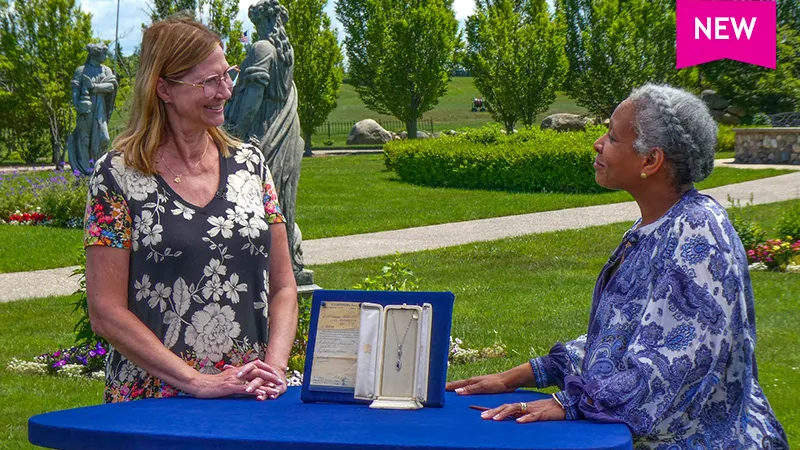 color photo of a lady discussing a necklace with appraiser Lea Koonce Ogundiran in front of sculptures and shrubery