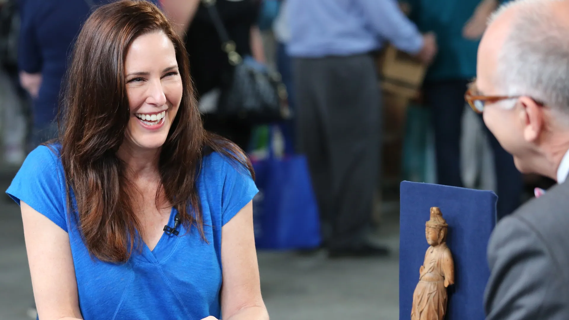 A seated female guest in a blue top smiles broadly while discussing an Asian artifact with appraiser Lark Mason.