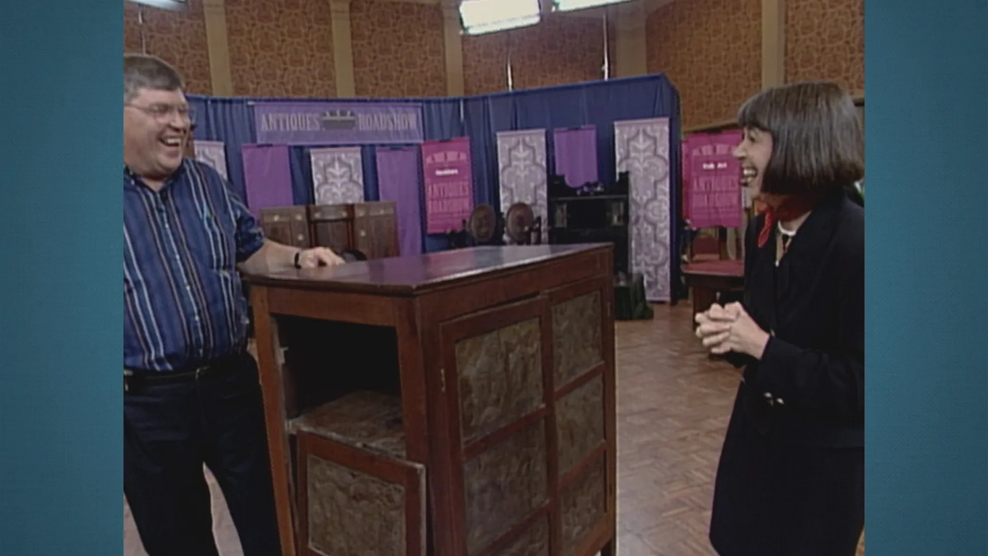 A man and woman smile while standing beside an antique chest of drawers