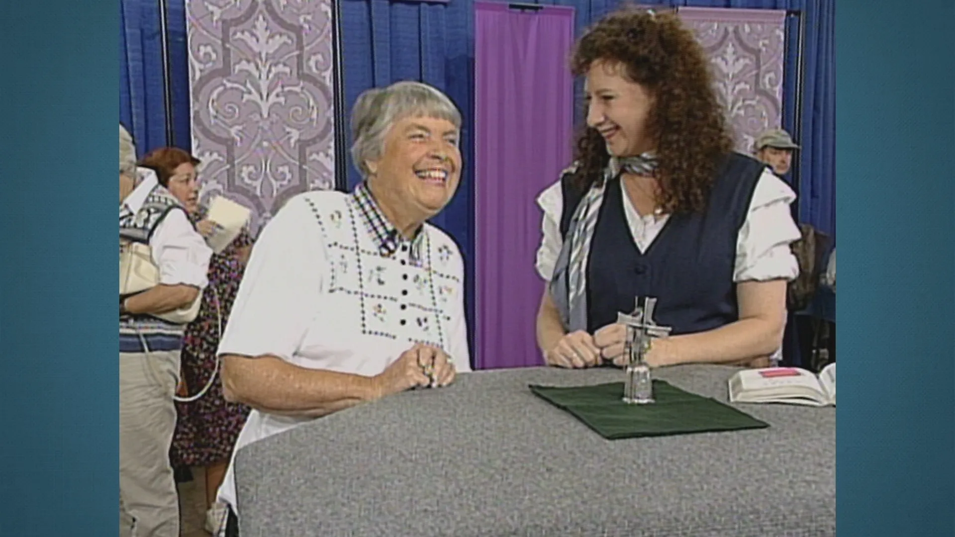 Two women standing and smiling, with a small antique silver windmill on a tabletop between them.