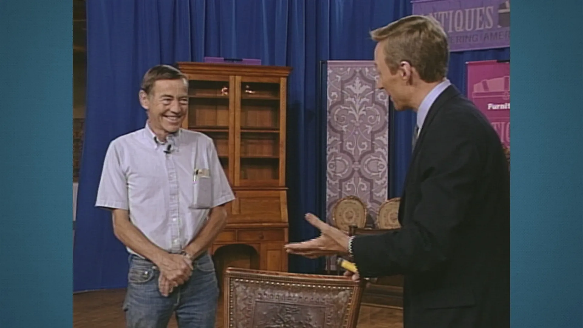 A man, standing, smiles while listening to appraiser Leslie Keno discuss an antique chair.