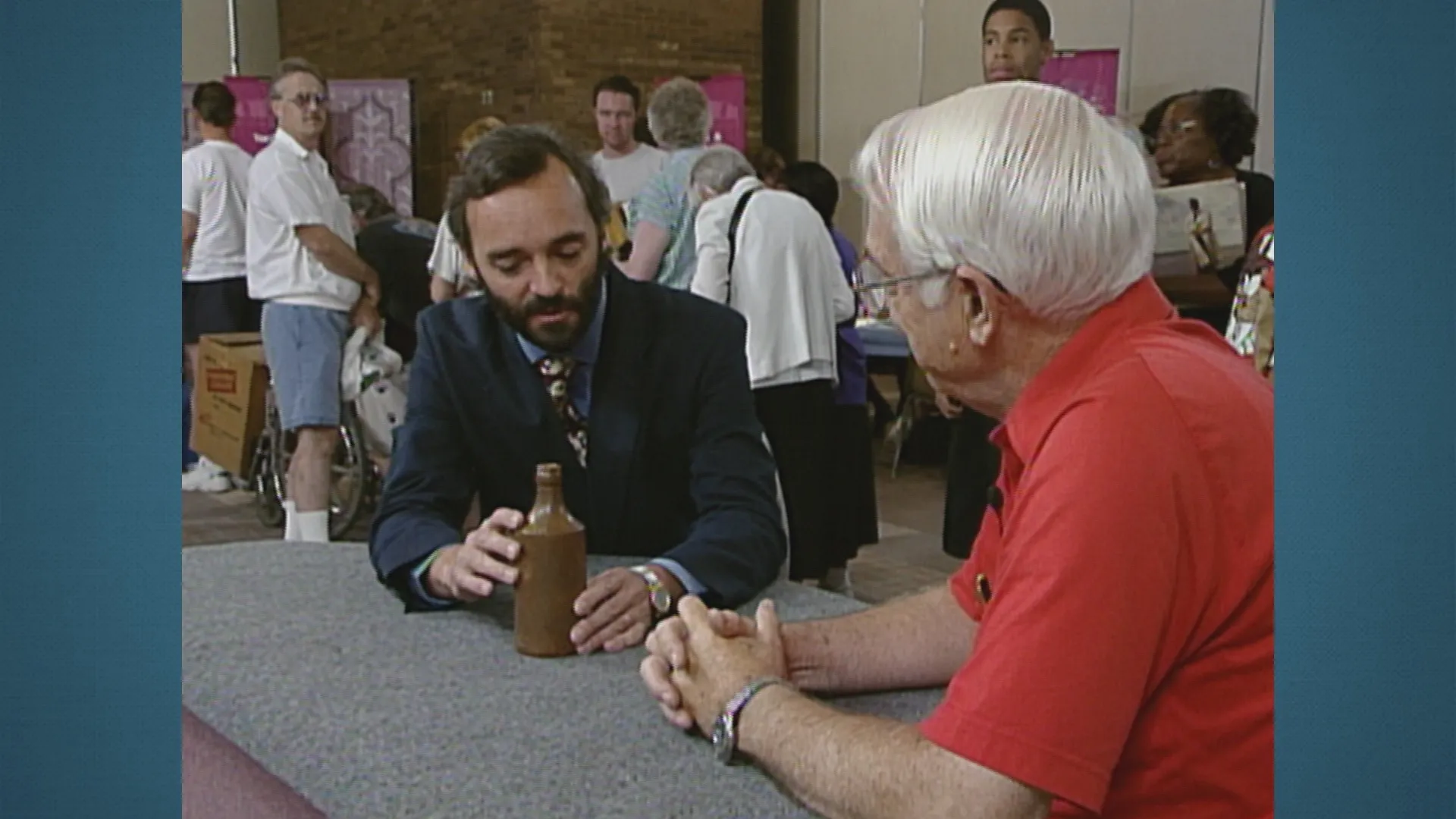 A man listens as appraiser Nick Dawes discusses a small antique earthenware jug while both are seated at a table.