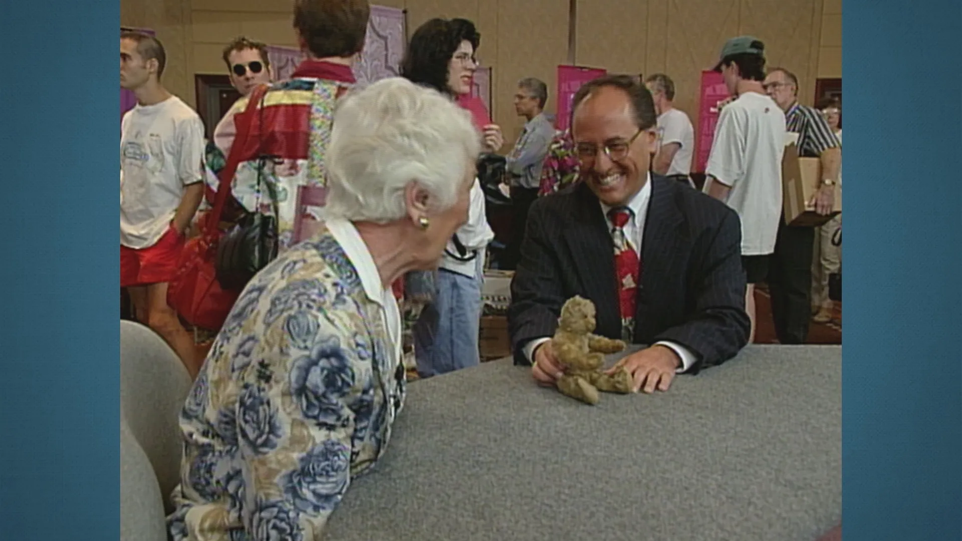 A man and woman, seated at a table, smile while discussing a small antique teddy bear.