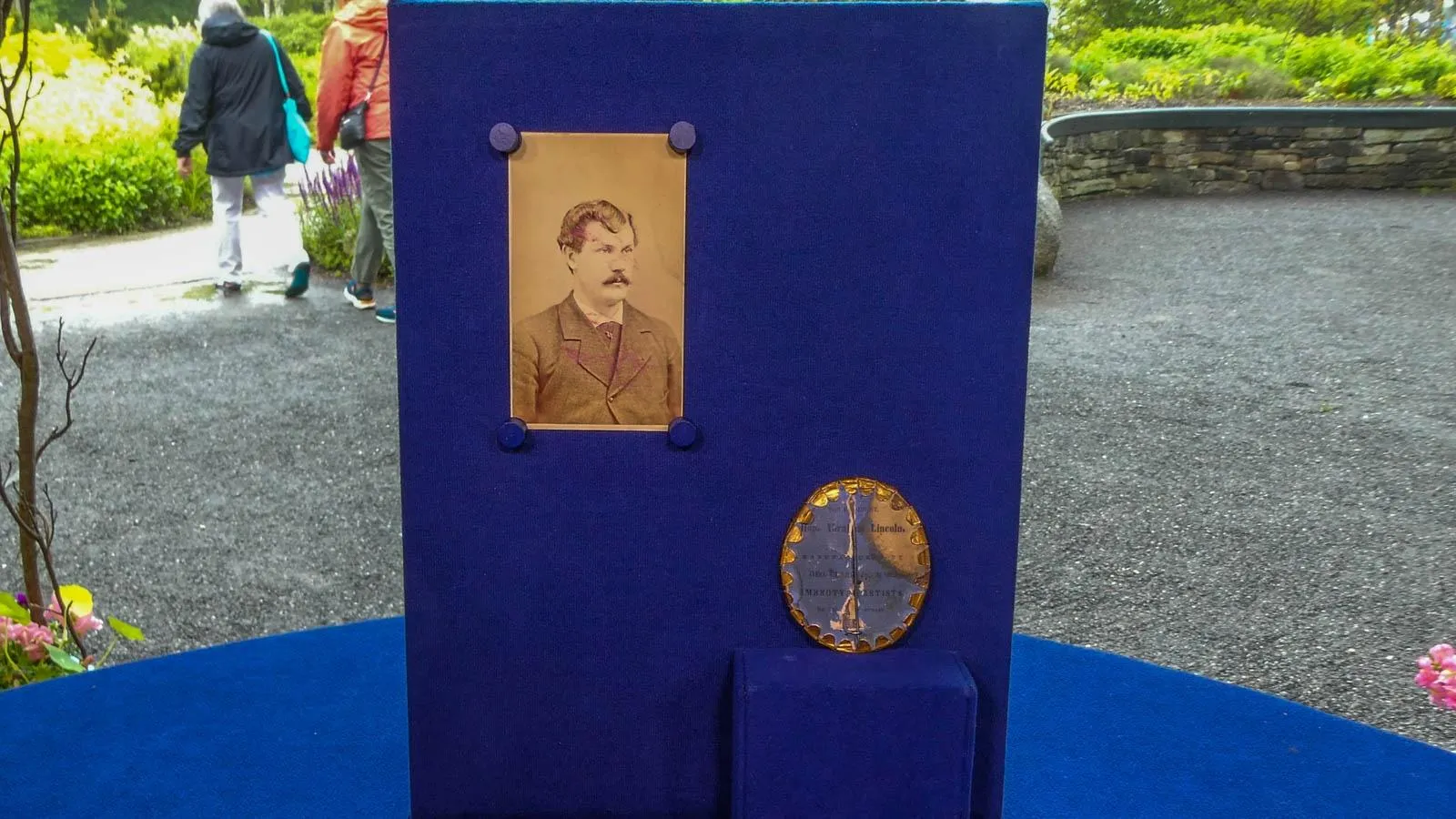 A yellowed photo of a man is mounted on a blue foam board. On the bottom left corner is a metallic campaign button propped on a blue display stand.