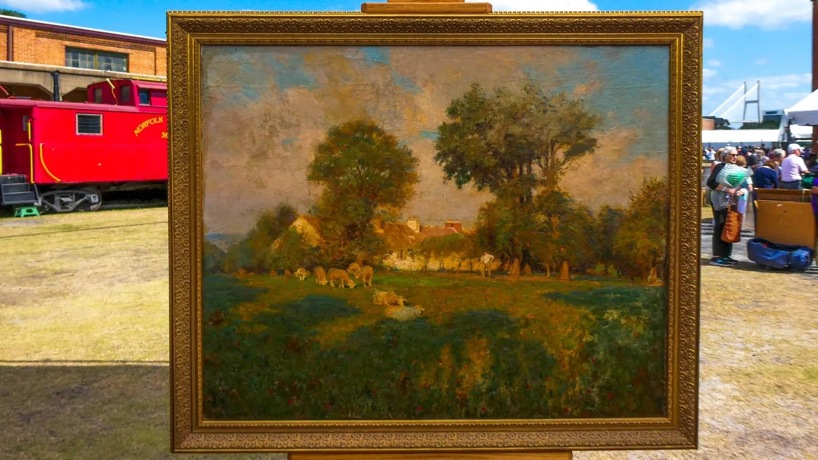 An oil painting of a farm landscape in a gold frame placed on an easel in an outdoor setting