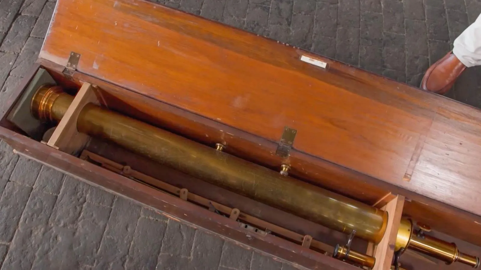 A gold telescope is placed in a wooden box  place on the ground on top of grey cobblestone