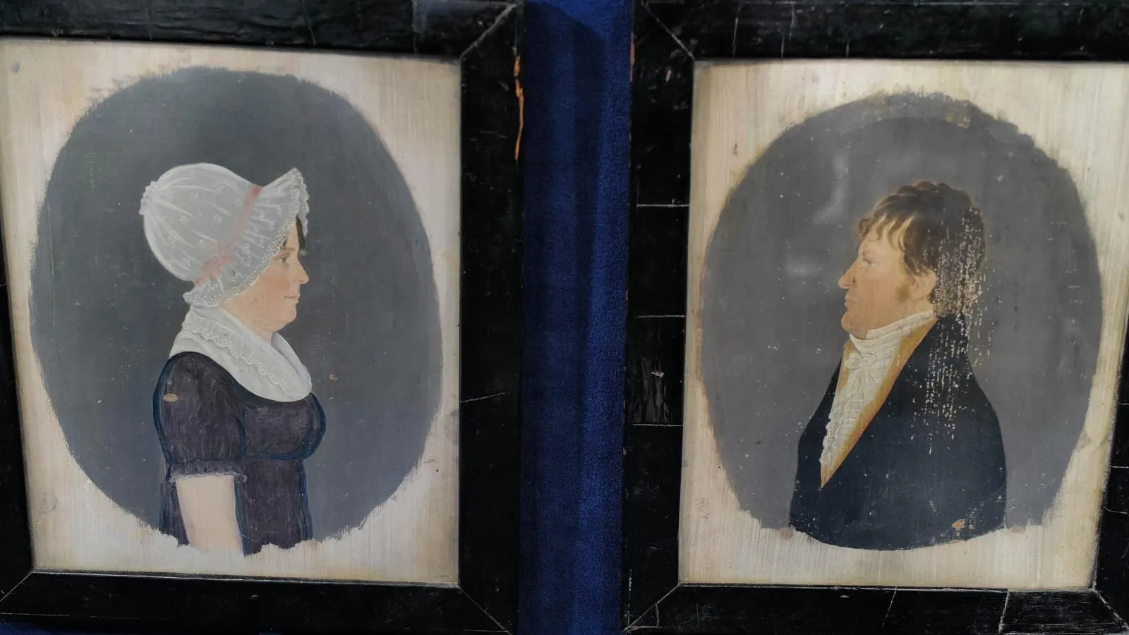 Two folk art hand painted portraits: a woman in a blue dress and white bonnet facing towards the right, and a man in a blue coat and white collard shirt facing his wife to the left.
