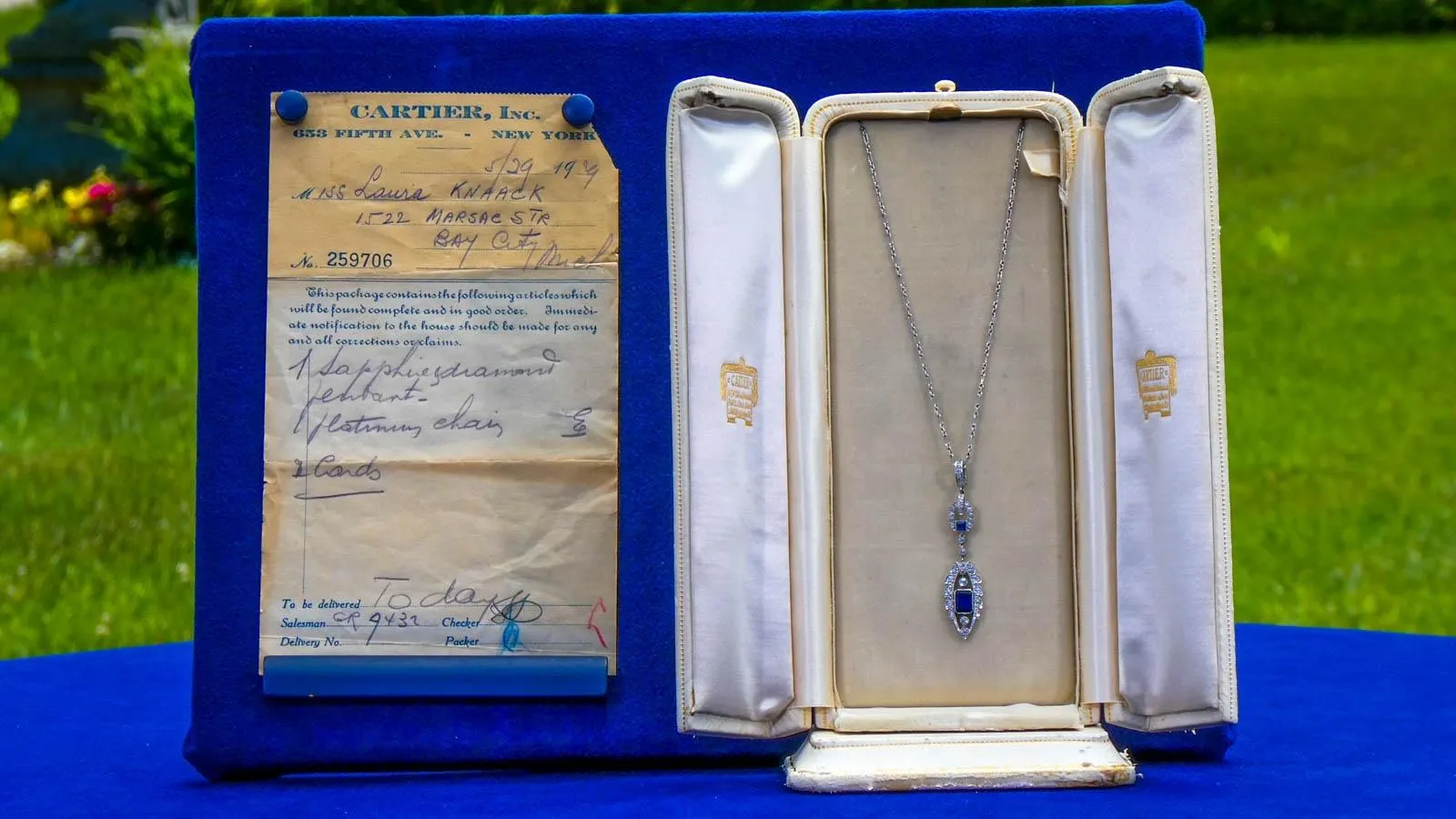 A Sapphire necklace eith platinum chain is in a display box with double doors on a table next to a paper cartier reciept.
