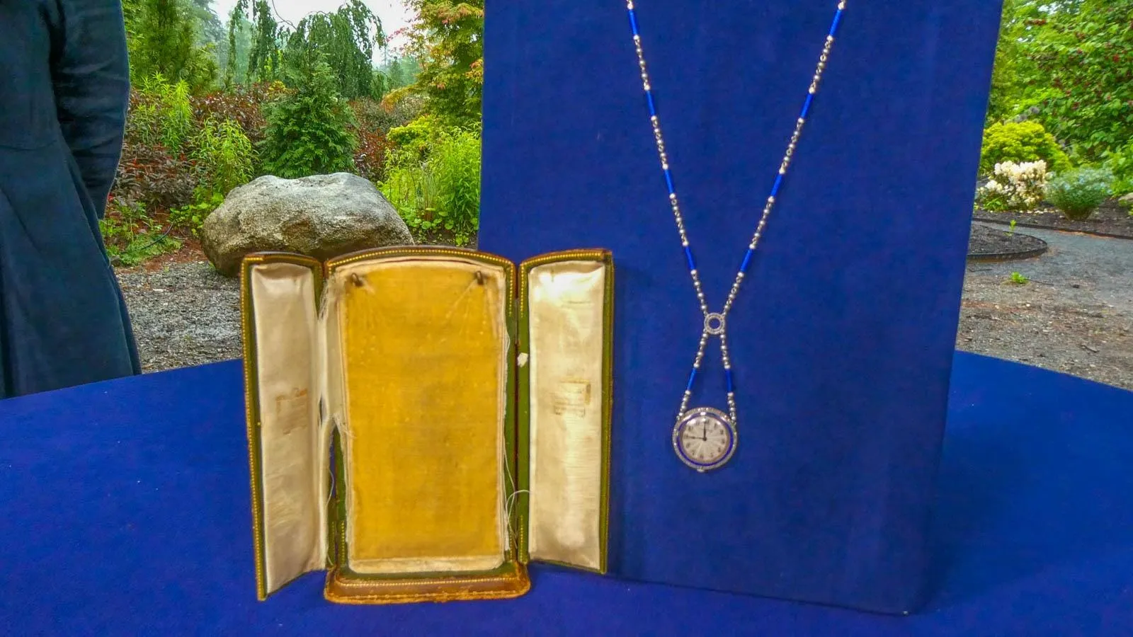 A pendant  watch with a silver and blue neck chain on a blue display board. Next to the necklace is the necklaces original box.