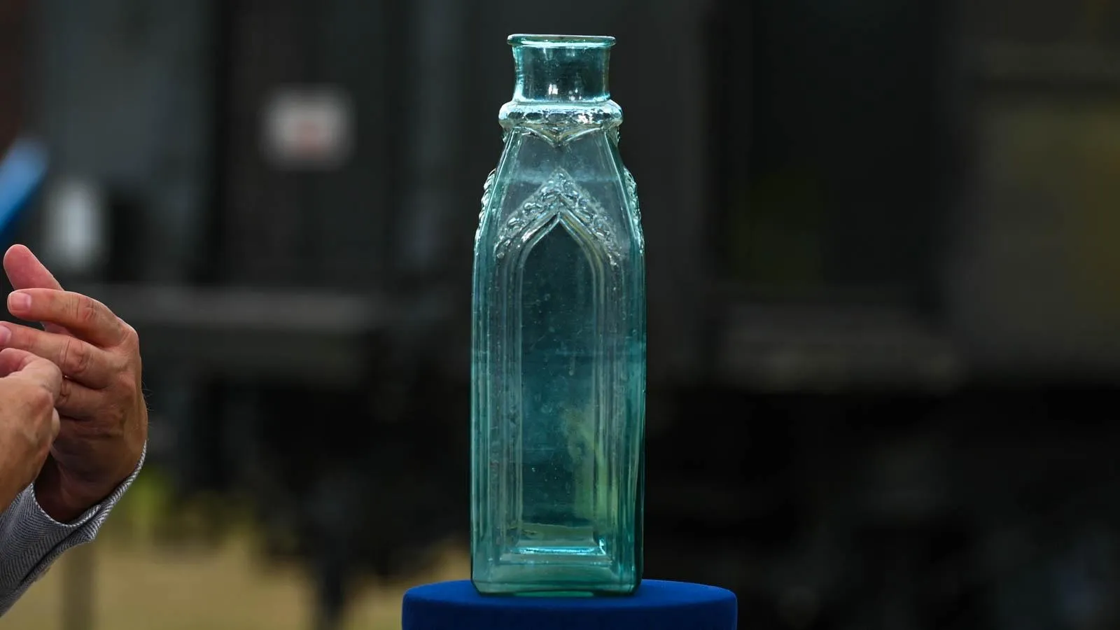 A glass bottle with intricate deatailing on the neck sits on top of a blue podium