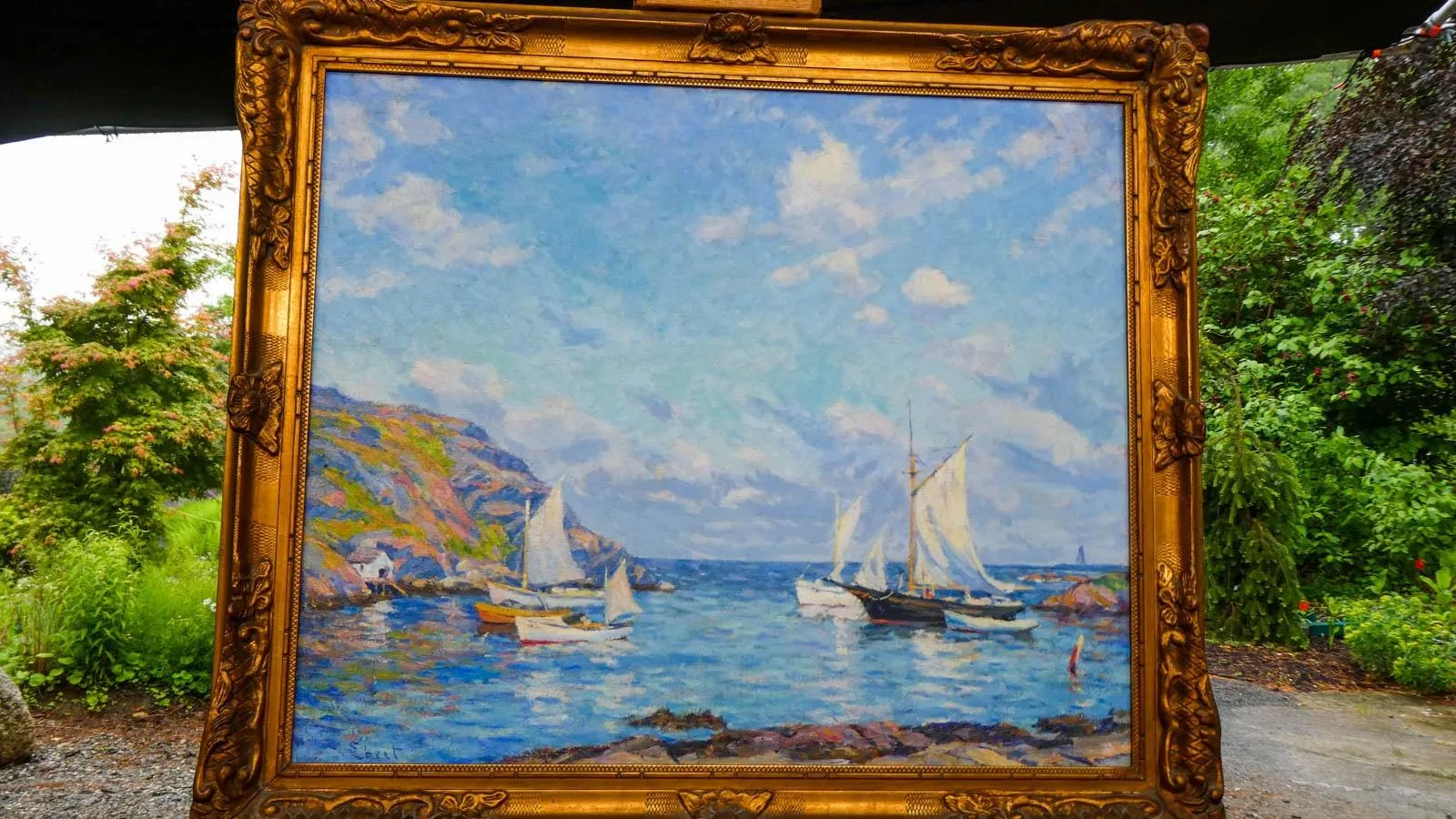 An oil painting of the ocean and sail boats is mounted in a gold ornate frame set up against a nature backdrop.