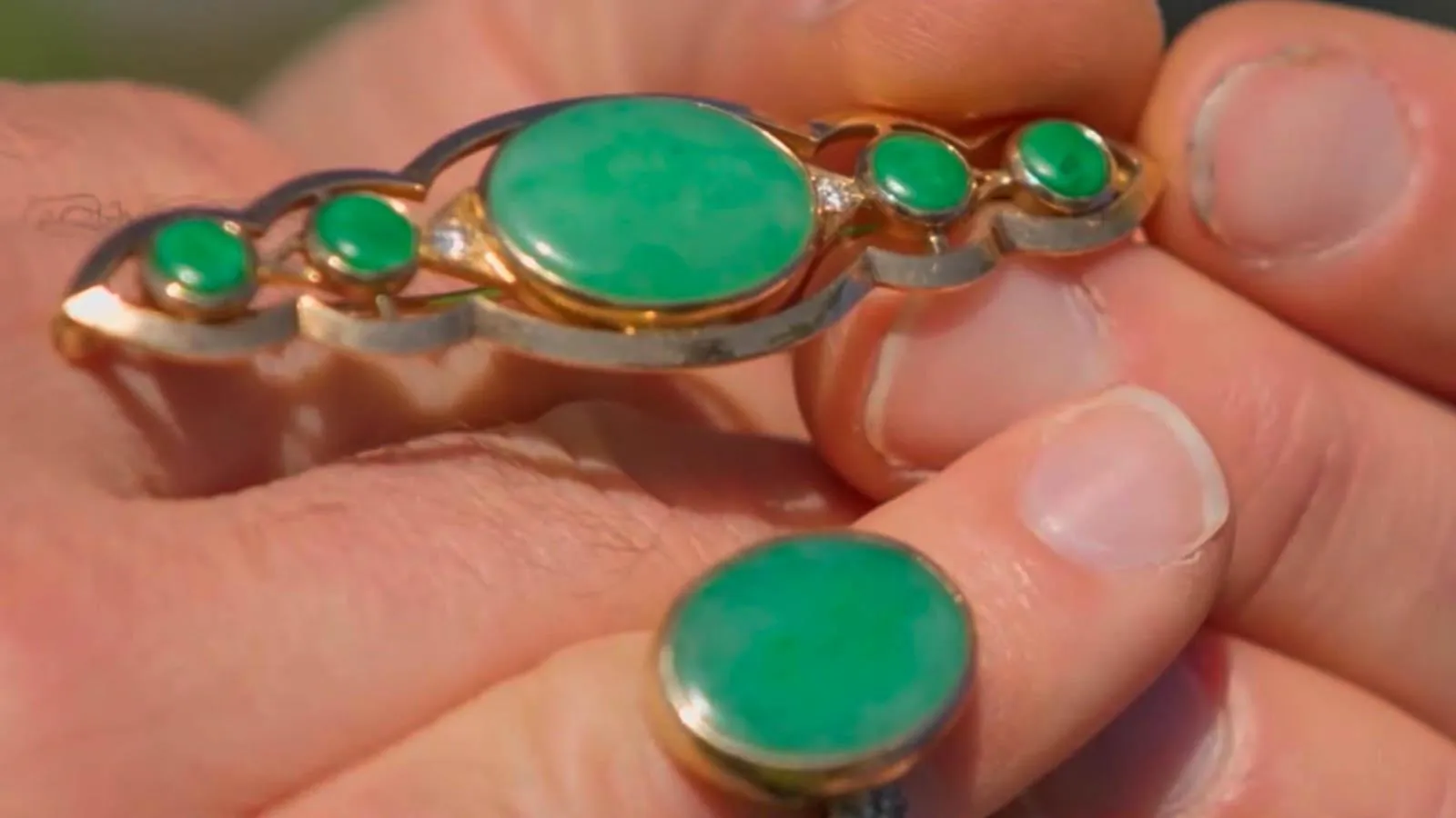 A close up of a hand holding a golden brooch adorned with green jadeite ovals. Next to the brooch on the display hand is a matching ring.