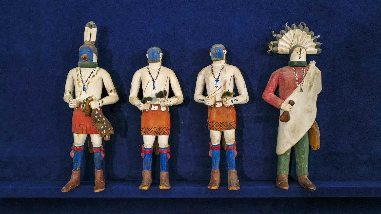 Four figures are aligned against a blue background. Three of the figures have blue facepaint and wear tribal skirts with the fourth having a headress with a sash across their chest.