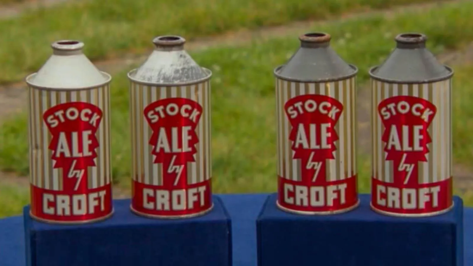 Four cone top cans with gold stripes and red writing sit in a line on top of a blue display stand.