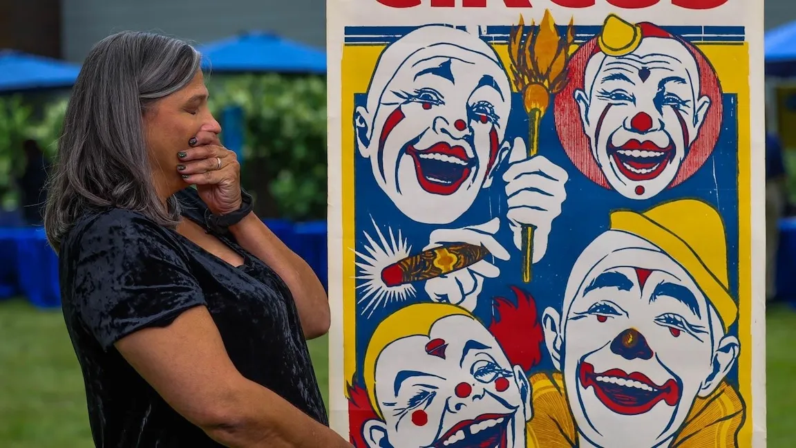 A woman in a black crushed velvet shirt looks shocked standing next to a paining of four different clown faces with colors of red, blue, and yellow.