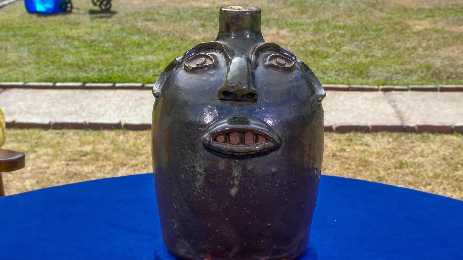 A black stoneware jug with a face sculpted onto it in a nueteral expression sits on top of a blue table.